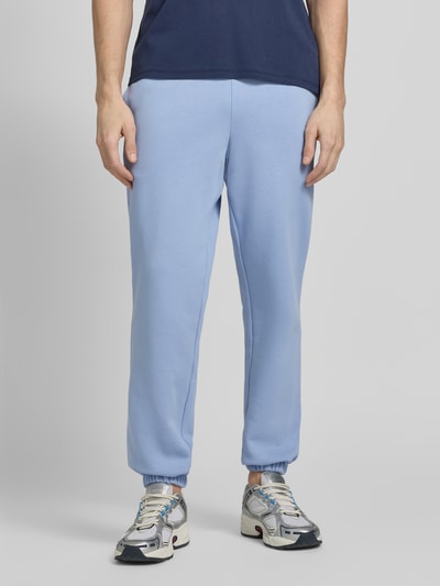Mężczyzna nosi Regular Fit Sweatpants aus Baumwoll-Mix od Lacoste, jasnoniebieski, logo, elastyczna talia i nogawki, bawełniany materiał, gładka faktura, boczne kieszenie, widok całej sylwetki z przodu.