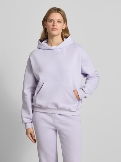 Kobiecy model nosi bluzę z kapturem z kieszenią kangurka model 'Iconic' od Gina Tricot, mieszanka bawełny, krój relaxed fit, jasnofioletowy kolor, gładka tekstura, całkowity widok z przodu.