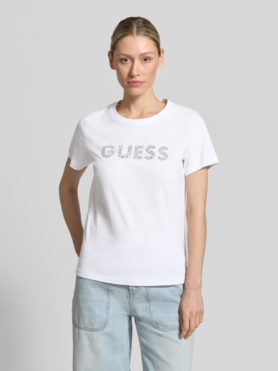 Frauenmodel trägt T-Shirt mit Label-Print und Strasssteinbesatz von Guess aus reiner Baumwolle, Straight Fit, Rundhalsausschnitt, Rückenlänge bei Größe S 59 cm, dezentes Weiß und helle Jeans, glatte Textur, Ganzkörperansicht von vorne.