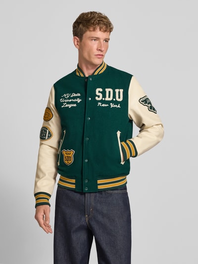 Männermodel trägt College-Jacke aus Woll-Mix mit Eingrifftaschen von Superdry in Grün und Creme, Regular Fit, Ganzkörperansicht von vorne.