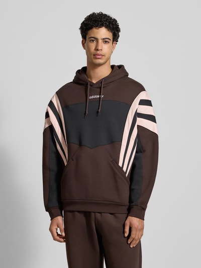 Männermodel trägt Hoodie mit Kapuze von adidas Originals in Braun mit schwarzen und beigen Details, Label-Stitching, Känguru-Tasche, Lockere-Schnitt, Kapuze mit Tunnelzug, Gerippte-Abschlüsse, Rückenlänge 71 cm, Ärmellänge 66 cm, Ganzkörperansicht von vorne.