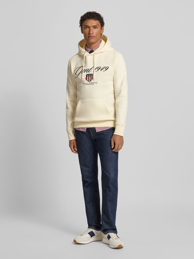 Mannermodel draagt Hoodie met Labelstitching en kangoeroezak van Gant in katoenmix, regular fit, met blauwe jeans, Ganzkörperansicht von vorne. Ruglengte maat M 70 cm, mouwlengte maat M 63 cm.