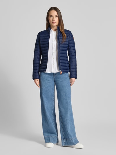 Frauenmodel trägt Slim Fit Steppjacke mit gefüttertem Stehkragen Modell 'Carly' von SAVE THE DUCK in Dunkelblau, Stepp-Optik, Ganzkörperansicht von vorne, Damenjacke mit Zweiwegereißverschluss und Reißverschlusstaschen, lockere Jeans und weißen Sneakern, ruhiger Hintergrund.