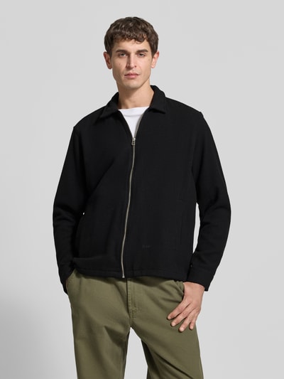 Męski model nosi Wolljacke aus Woll-Mix od Forét, czarny kolor, krój regular fit, kołnierzyk, zamek błyskawiczny, kieszenie, długość pleców w rozmiarze M 71 cm, szerokość ramion w rozmiarze M 47 cm, długość rękawa w rozmiarze M 67 cm, widok całej sylwetki z przodu.