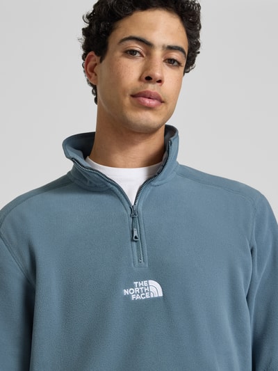 Męski model nosi Fleecepullover mit Reißverschluss Modell 'GLACIER' von The North Face w kolorze niebieskoszarym, krój regular fit, stójka, label-stitching, zamek błyskawiczny, długość tyłu w rozmiarze M: 70 cm, długość rękawa w rozmiarze M: 67 cm, tekstura miękkiego polaru, zbliżenie na górną część ciała.