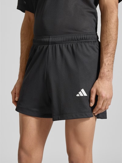 Mężczyzna nosi Straight Fit Shorts mit Logo-Print in Mesh-Optik od adidas Training, czarne krótkie spodenki, mesh, elastyczny pas, szerokość w rozmiarze M 39 cm, długość nogawki 12 cm, widok na nogi i talię, zbliżenie na dolną część ciała
