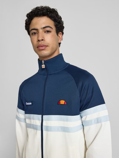 Męski model nosi bluza rozpinana z mieszanki bawełny, prosty krój, model 'RIMINI' od Ellesse, stójka, zamek błyskawiczny, detale z logo, elastyczne wykończenia, długość tyłu w rozmiarze M: 71 cm, długość rękawa w rozmiarze M: 43 cm, kolor granatowy i kremowy, struktura gładka, całkowite ujęcie przodu.