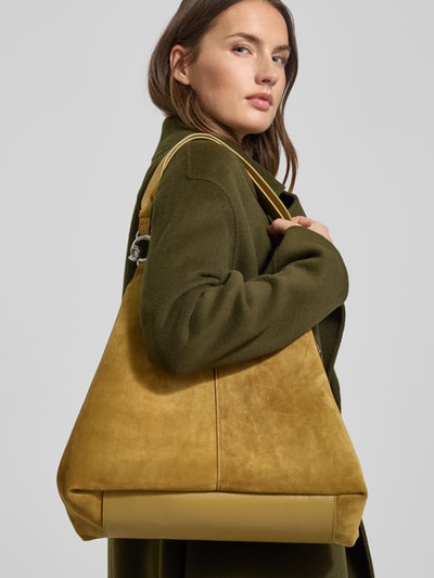 Kobiece modelka nosi torebkę z uchwytem Handtasche mit Tragehenkel od Coccinelle, zapięcie na magnes, detal z logo, szerokie 40 cm, długość 29 cm, głębokość 16 cm, pasek na ramię 52 cm, tekstura zamszowa, ciepły żółty kolor, półmatowe wykończenie, półzbliżenie z przodu.