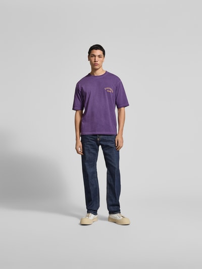 Männermodel trägt Loose Fit T-Shirt aus reiner Baumwolle von Dsquared2 in Lila mit Label-Print, Rundhalsausschnitt, Ganzkörperansicht von vorne. Herren T-Shirt, kurzärmelig, Rückenlänge bei Größe M: 68 cm, glatte Struktur, kombiniert mit Jeans und Sneakern.