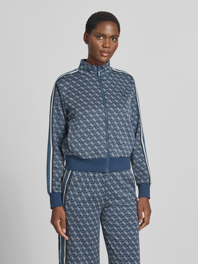 Frauenmodel trägt Regular Fit Sweatjacke mit Allover-Logo-Print von GUESS, blauer Ton mit weißem Muster, Reißverschluss, Nahaufnahme Oberkörper. Damen Sweatjacke, elastischer Material-Mix, Stehkragen, elastische Abschlüsse von GUESS Activewear.