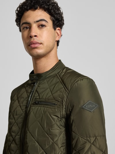 Männermodel trägt Regular Fit Jacke in Stepp-Optik von Replay in Olivgrün mit Stehkragen, Logo-Applikation, Nahaufnahme Oberkörper. Rückenlänge 66 cm, Schulterbreite 43 cm, Brustweite 52 cm, Ärmellänge 63 cm, Brusttasche und Eingrifftaschen mit Reißverschluss.