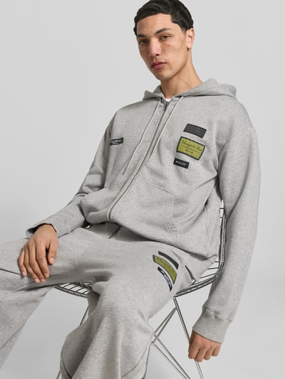 Männermodel trägt Regular Fit Sweatjacke mit Logo-Patch von MM6 Maison Margiela in Grau, reine Baumwolle, Nahaufnahme Oberkörper. Rückenlänge 69 cm, Schulterbreite 45 cm, Ärmellänge 72 cm, Kapuze mit Tunnelzug, Reißverschluss.