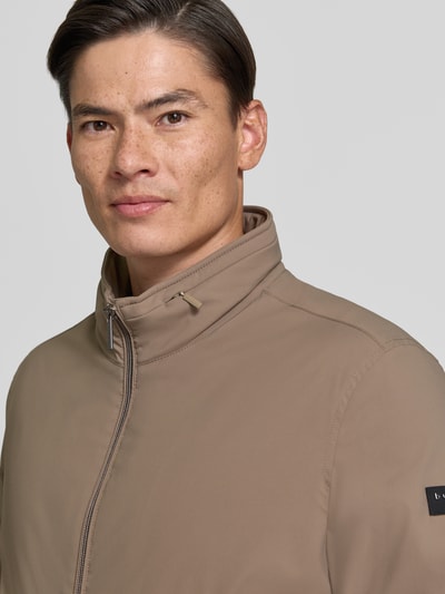 Männermodel trägt Regular Fit Jacke mit Stójka von bugatti w kolorze beżowym, zamek błyskawiczny, wpuszczane kieszenie, krój regular fit, elastyczny pas, długość tyłu w rozmiarze 50: 67 cm, szerokość ramion w rozmiarze 50: 45 cm, szerokość klatki piersiowej w rozmiarze 50: 58 cm, długość rękawów w rozmiarze 50: 67 cm, zbliżenie na górną część tułowia