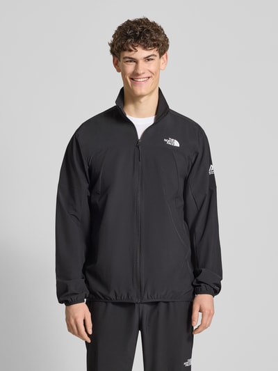 Męski model nosi Regular Fit Jacke mit Stehkragen Modell 'Mountain' von The North Face, czarna elastyczna mieszanka materiałów, nadruk z logo, zamek błyskawiczny, wpuszczane kieszenie, elastyczne wykończenia, długość tyłu w rozmiarze M 73 cm, szerokość ramion w rozmiarze M 43 cm, szerokość klatki piersiowej w rozmiarze M 56 cm, długość rękawów w rozmiarze M 70 cm, krój regular fit, całkowity widok z przodu