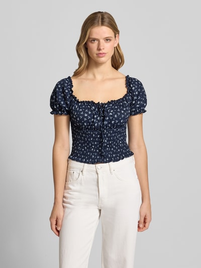 Frauenmodel trägt Slim Fit Blusentop mit Allover-Muster Modell 'HILLARY' von Only in floraler Optik, Cropped-Länge, Henley-Ausschnitt, Kurzarm mit Raffungen, dunkelblauer Grundton, feine Musterstruktur, Ganzkörperansicht von vorne.