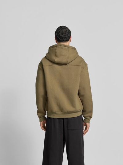Männermodel trägt Oversized Sweatjacke aus Baumwolle von RHUDE in Olive mit Label-Stitching, elastischen Abschlüssen, Rückenlänge 70 cm, Ganzkörperansicht Rückansicht.