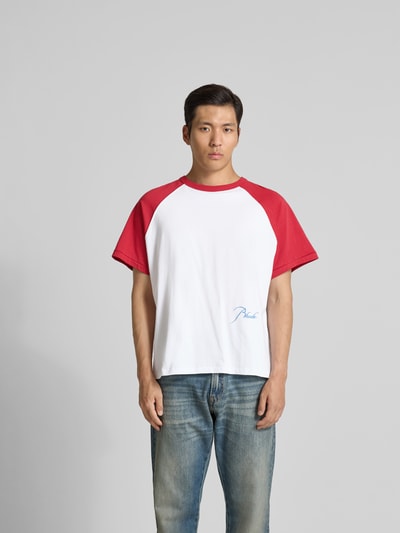 Männermodel trägt T-Shirt mit Label-Stitching von RHUDE in Weiß mit roten Ärmeln aus reiner Baumwolle, gerade Passform, Rundhalsausschnitt, kurzärmelig, Rückenlänge bei Größe M 68 cm, Ganzkörperansicht von vorne.