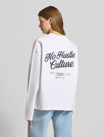Frauenmodel trägt Oversized Longsleeve mit rückseitigem Statement-Print von Karo Kauer, bawełna, krój oversized fit, okrągły dekolt, nadruk z logo, długość tyłu w rozmiarze S: 65 cm, jasne kolory, proste formy, widoczna tekstura materiału. Ganzkörperansicht Rückansicht.