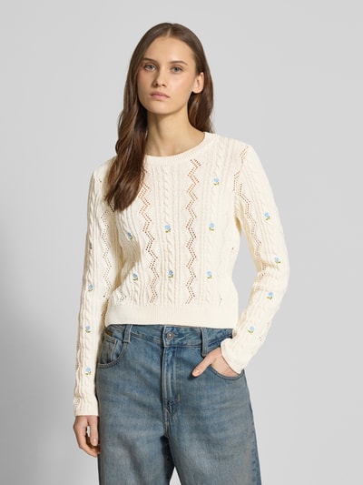 Modelka kobieca ma na sobie Regular Fit Strickpullover aus recyceltem Baumwoll-Mix Modell 'RITA LIFE' od Only, kremowy sweter z ażurowym i warkoczowym wzorem, haftowane aplikacje, okrągły dekolt, długie rękawy, widoczne prążkowane ściągacze, przędza bawełniana z recyklingu, krój regular fit, widok na górną część ciała z przodu.