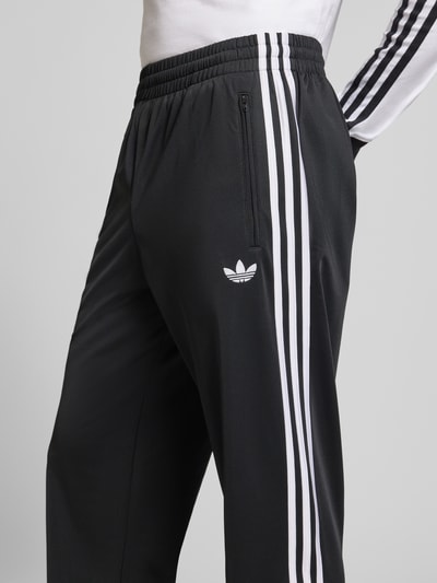 Männermodel trägt Sweatpants mit elastischem Bund von adidas Originals in Schwarz mit weißen Streifen, Nahaufnahme Oberschenkel, lockerer Schnitt, Label-Stitching und Reißverschlusstaschen.