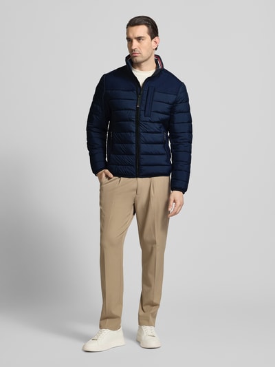 Męski model nosi Regular Fit Steppjacke mit seitlichen Stretcheinsätzen von Tom Tailor, granatowa, pikowana, detal logo, krój regular fit, stójka, zamek, elastyczne wykończenia, wodoodporna, kieszenie; całopostaciowy widok z przodu