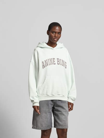 Frauenmodel trägt Oversized Hoodie mit Logo-Stitching von Anine Bing in Mintgrün aus reiner Baumwolle mit Kapuze und Känguru-Tasche, Oversized-Fit, Rückenlänge bei Größe S 64 cm, Ganzkörperansicht von vorne.