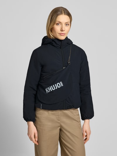 Vrouwenmodel draagt zwarte jack met capuchon en labeldetail, regular fit model 'SHELLY2' van khujo, vooraanzicht, ruglengte maat S 56 cm, schouderbreedte 52 cm, borstwijdte 46 cm, mouwlengte 52 cm. Nahaufnahme Oberkörper