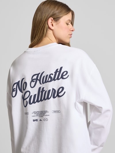Modelka nosi Oversized Longsleeve mit rückseitigem Statement-Print von Karo Kauer, biała bawełna, krój oversized fit, okrągły dekolt, nadruk z logo, długość tyłu w rozmiarze S: 65 cm, nadruk z napisem na plecach. Ujęcie: z tyłu, górna część ciała.
