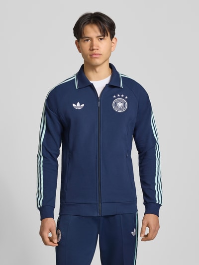 Männermodel trägt Regular Fit Trainingsjacke mit Deutschland Logo von adidas Sportswear in Dunkelblau, unifarben, Kontraststreifen, Ganzkörperansicht von vorne.