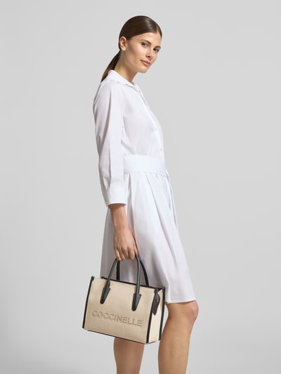 Frauenmodel trägt helle Handtasche mit Label-Stitching von Coccinelle, Hauptfach mit Reißverschluss, Tragehenkel, Schulterriemen, Breite 27 cm, Länge 20 cm, Tiefe 13 cm, Länge Schulterriemen 105 cm; Ganzkörperansicht von vorne.