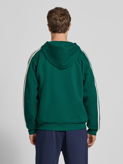 Männermodel trägt Regular Fit Sweatjacke aus Baumwoll-Mix von adidas, grün mit weißen Streifen, Herren Sweatjacke von adidas Sportswear, Ganzkörperansicht Rückansicht.
