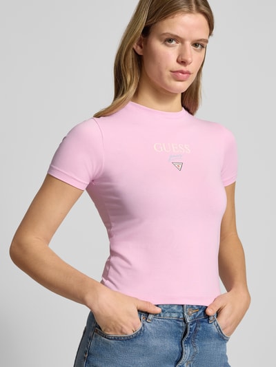 Vrouwenmodel draagt T-shirt met logo en ronde hals van Guess, mix van katoen en elastaan, slim fit, lichtroze kleur, detailopname bovenlichaam.