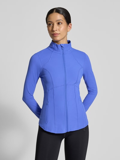 Frauenmodel trägt Slim Fit Jacke mit Stehkragen Modell 'Storm' von Fabletics in Blau, Reißverschluss, Daumenschlaufen, Ganzkörperansicht von vorne.