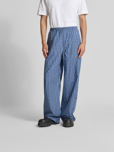 Męski model nosi Pyjama-Hose aus reiner Baumwolle von VETEMENTS w niebieską kratkę, krój relaxed fit, elastyczny pas, wpuszczane kieszenie, detal z logo, długość wewnętrzna nogawki 86 cm, szerokość w rozmiarze M: 46 cm, widok całej sylwetki z przodu.