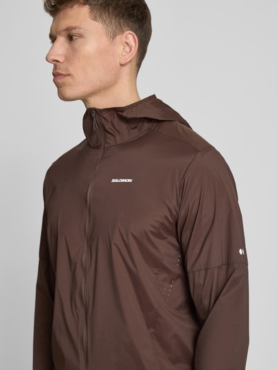 Männermodel trägt Windbreaker mit Kapuze und Logo Modell 'SENSE AERO' von Salomon in Braun, Material-Mix mit Stretch-Anteil, Regular-Fit, Nahaufnahme Oberkörper. Rückenlänge 71 cm, Schulterbreite 44 cm, Brustweite 41 cm, Ärmellänge 67 cm.