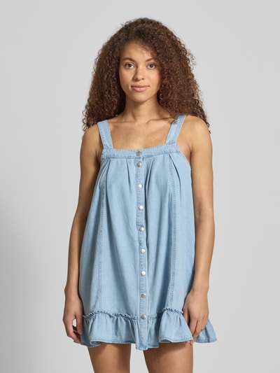 Frauenmodel trägt Jeanskleid aus reiner Baumwolle Modell 'OREGON' von Noisy May in Hellblau mit Rüschen und Druckknöpfen, Ganzkörperansicht von vorne.