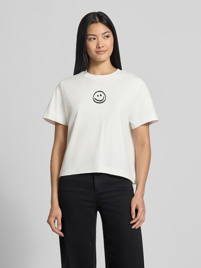 Kobiece model prezentuje damski t-shirt Boxy Fit T-Shirt aus reiner Baumwolle Model 'SARA' od VERO MODA, biały z czarnym nadrukiem, gładka tekstura, całkowity widok z przodu