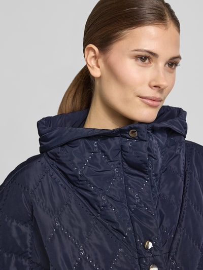 Frauenmodel trägt Cape mit Steppnähten und Eingrifftaschen von Copenhagen Muse in Dunkelblau mit Knopfleiste, Nahaufnahme Oberkörper. Rückenlänge bei Größe S/M: 66 cm, Schulterbreite: 42 cm, Brustweite: 25 cm, Relaxed-Fit, hochgeschlossener Kragen.