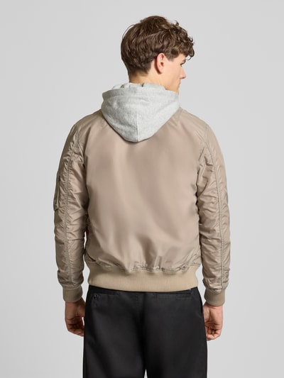 Männermodel trägt Blouson mit Logo-Anhänger Modell 'MA-1 TT' von Alpha Industries in Beige, Kapuze grau, lockere Passform, Ganzkörperansicht Rückansicht. Rückenlänge 69 cm, Schulterbreite 46 cm, Brustweite 54 cm, Ärmellänge 67 cm, elastische Abschlüsse, Patch und Reißverschluss sichtbar.