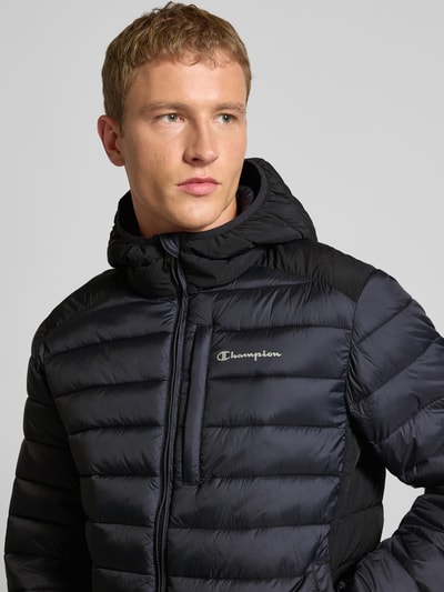 Männermodel trägt Steppjacke mit Kapuze und Logo von CHAMPION in Schwarz, Reißverschluss, Eingrifftaschen, Nahaufnahme Oberkörper. Rückenlänge 71 cm, Schulterbreite 51 cm, Brustweite 61 cm, Ärmellänge 69 cm. Regular-Fit.