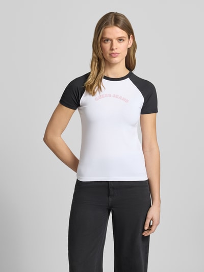 Vrouwenmodel draagt Slim Fit T-shirt met ronde hals en logo van Guess Jeans in wit met zwarte mouwen, mix van katoen en elastaan, ruglengte maat S 53 cm, slim fit, logoprint, ganzekörpervooraanzicht.