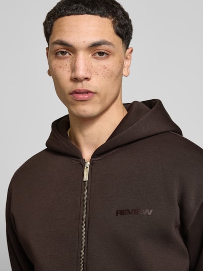 Männermodel trägt Sweatjacke mit Känguru-Tasche von REVIEW aus Baumwoll-Mix im Relaxed-Fit, braun, Reißverschluss, Nahaufnahme Oberkörper.