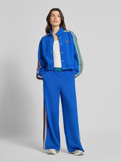 Frauenmodel trägt Sweatjacke mit Kontraststreifen Modell 'Brooklynn' von Lollys Laundry, Material-Mix mit Stretch-Anteil, Relaxed-Fit, Stehkragen, Knopfleiste, leuchtend blau mit farbigen Streifen und Stitching, Ganzkörperansicht von vorne.