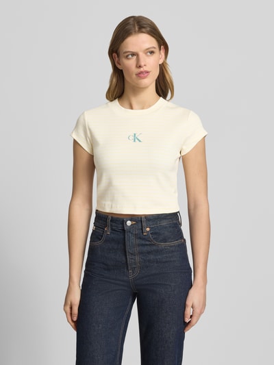 Modelka nosi Cropped T-Shirt mit Streifenmuster und Logo od Calvin Klein Jeans, kremowe tony, okrągły dekolt, logo z przodu, bawełna z domieszką elastanu, wzór w paski, długość tyłu w rozmiarze S: 39 cm, widok całej sylwetki z przodu