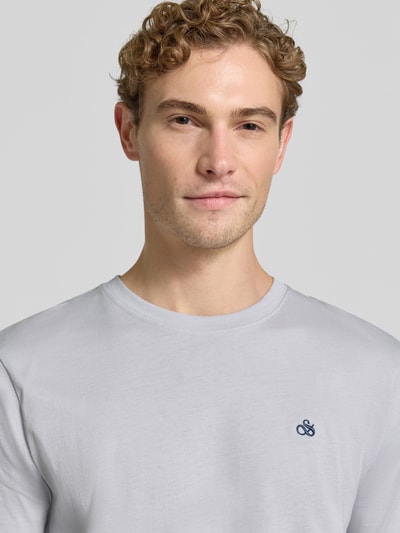 Męski model nosi t-shirt aus reiner Baumwolle von Scotch & Soda, jasnoszary kolor, wyhaftowane logo, luźny krój, okrągły dekolt, długość tyłu w rozmiarze M: 70 cm, zbliżenie na górną część ciała.