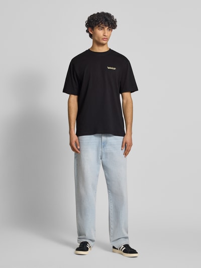Männermodel trägt ein unifarbenes, schwarzes Relaxed Fit T-Shirt aus reiner Baumwolle Modell 'FRED DU MARTIN' von Only & Sons mit Motiv-Print, Rundhals und kurzen Ärmeln, feine Rippenstruktur, Ganzkörperansicht von vorne.