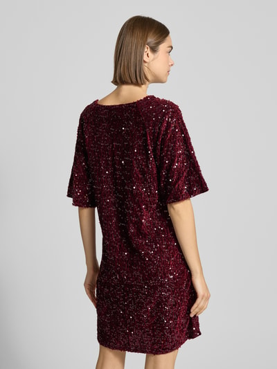 Kobieta nosi sukienkę koktajlową z cekinowym obszyciem Schmales Cocktailkleid mit Paillettenbesatz Modell 'KAM' od Pieces, efekt: jednolity kolor, efekt błyszczący, krój: wąski, długość mini, dekolt w łódkę, aplikacja: wyhaftowane logo, rękaw: krótki, prosty dół, wykończenie materiału: aksamit; zdjęcie całej sylwetki z tyłu.