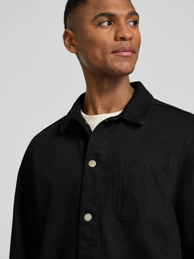 Männermodel trägt Regular Fit Overshirt mit Brusttasche Modell 'Layton' von Les Deux aus Reine Baumwolle in Schwarz, Nahaufnahme Oberkörper. Kentkragen, Knopfleiste, Eingrifftaschen, Sportmanschetten, Rückenlänge bei Größe M: 67 cm.