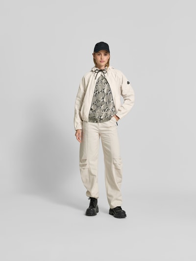 Frauenmodel trägt Jacke mit Zweiwege-Reißverschluss von Moose Knuckles in Beige mit Kapuze, Eingrifftaschen, Relaxed-Fit, Ganzkörperansicht von vorne. Rückenlänge 56 cm, Brustweite 56 cm, Ärmellänge 44 cm, Größe S.