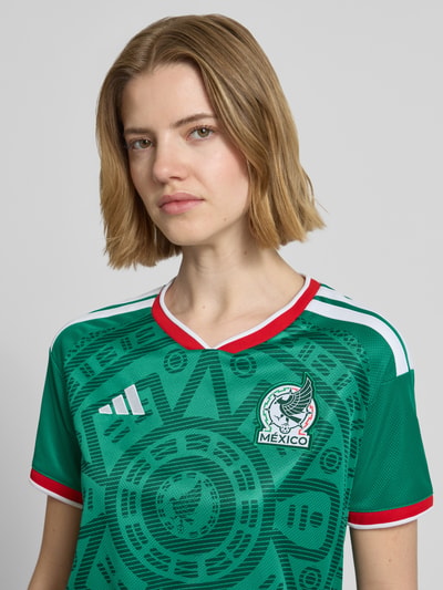Frauenmodel trägt Mexiko Heimtrikot 2026 von adidas Sportswear, Optik: Mehrfarbig, Passform: Regular-Fit, FMF-Applikation, Kontraststreifen, Logo-Stitching, V-Ausschnitt, Ärmelform: Kurzarm, Nahaufnahme Oberkörper.